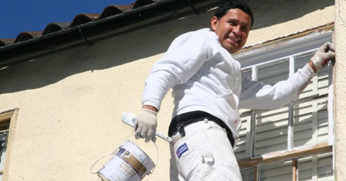 A smiling painter in a white uniform stands on a ladder against a stucco house, painting window trim and holding a paint can and brush.