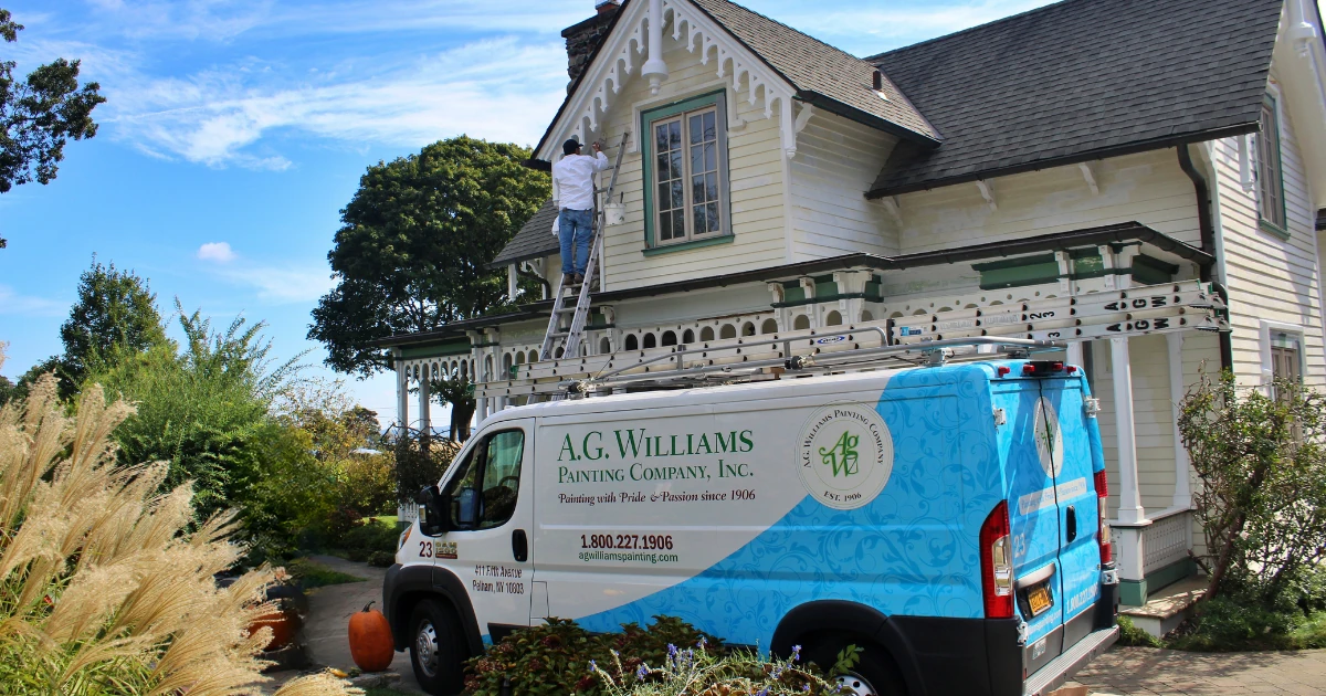 An A.G. Williams Painting Company branded van parked in front of a light-yellow Victorian house while a painter on a ladder works on the upper trim.