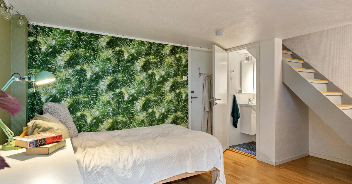 Bedroom with a green tropical-leaf wallpaper accent wall, a single bed, a desk with books, a staircase leading to an upper floor, and an open door to a small bathroom.