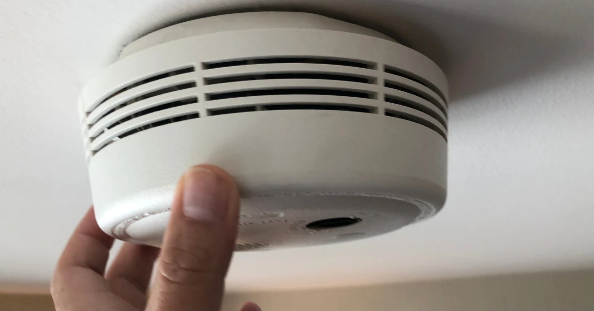 Close-up of a hand touching a white smoke or carbon monoxide alarm mounted on a ceiling.