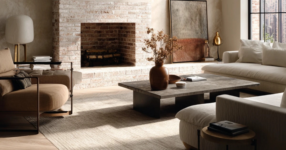 A living room with a whitewashed brick fireplace, plastered walls, neutral sofas, and a stone coffee table with a dried floral arrangement.