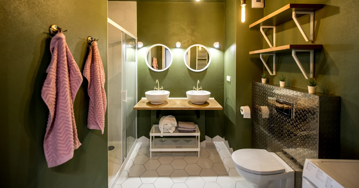 An olive-green bathroom with two round mirrors above twin vessel sinks on a wooden counter, hexagonal white floor tile, pink towels, and wood shelving.