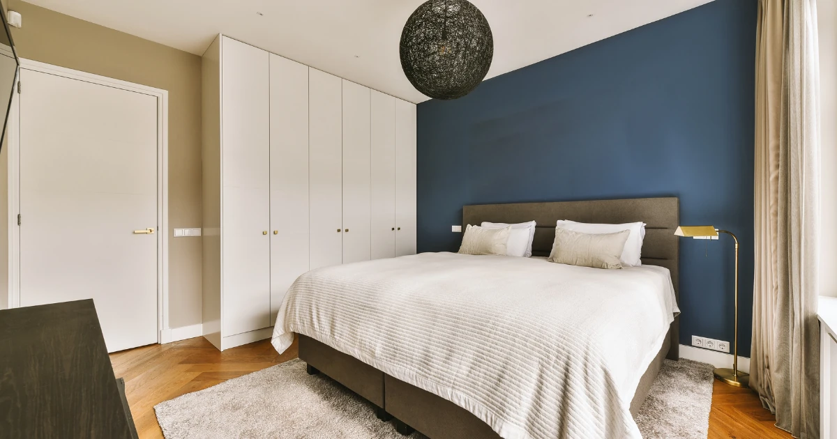 Bedroom with a navy blue painted accent wall behind an upholstered bed, white built-in wardrobes, and a black woven pendant light.