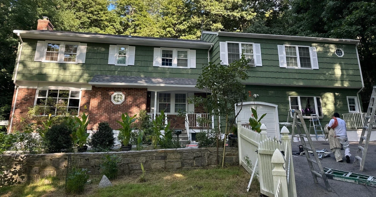 A green two-story colonial house with brick accents and white shutters undergoing exterior painting, with a worker and multiple ladders set up in the driveway.