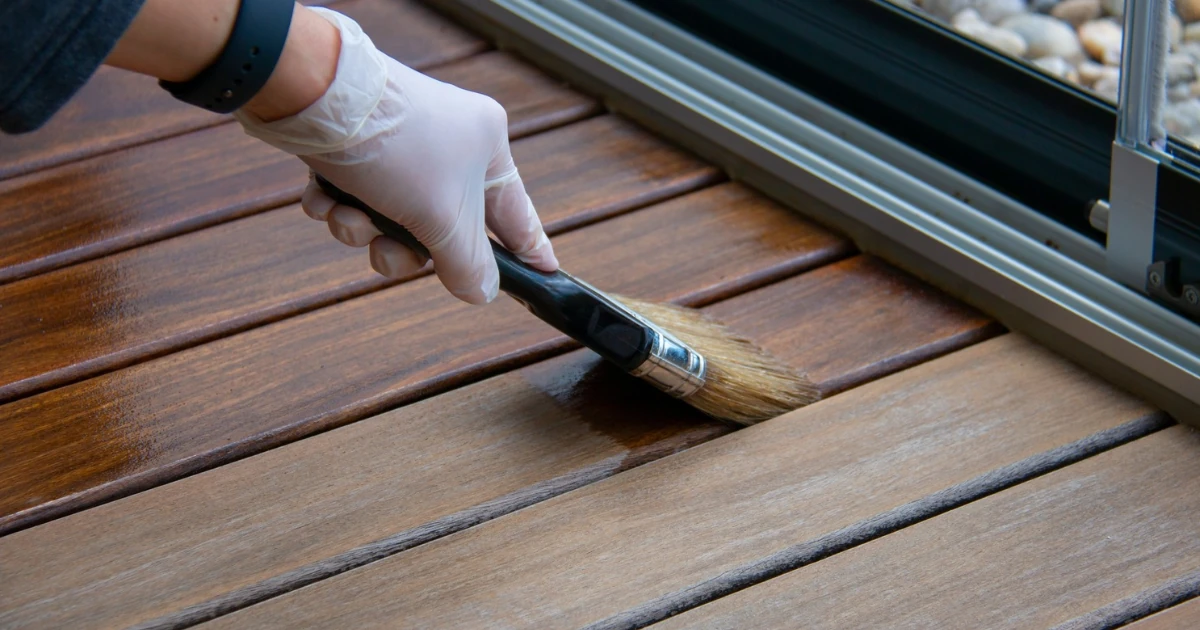 Close-up of a gloved hand using a paintbrush to apply wood stain to deck boards beside a sliding door track.