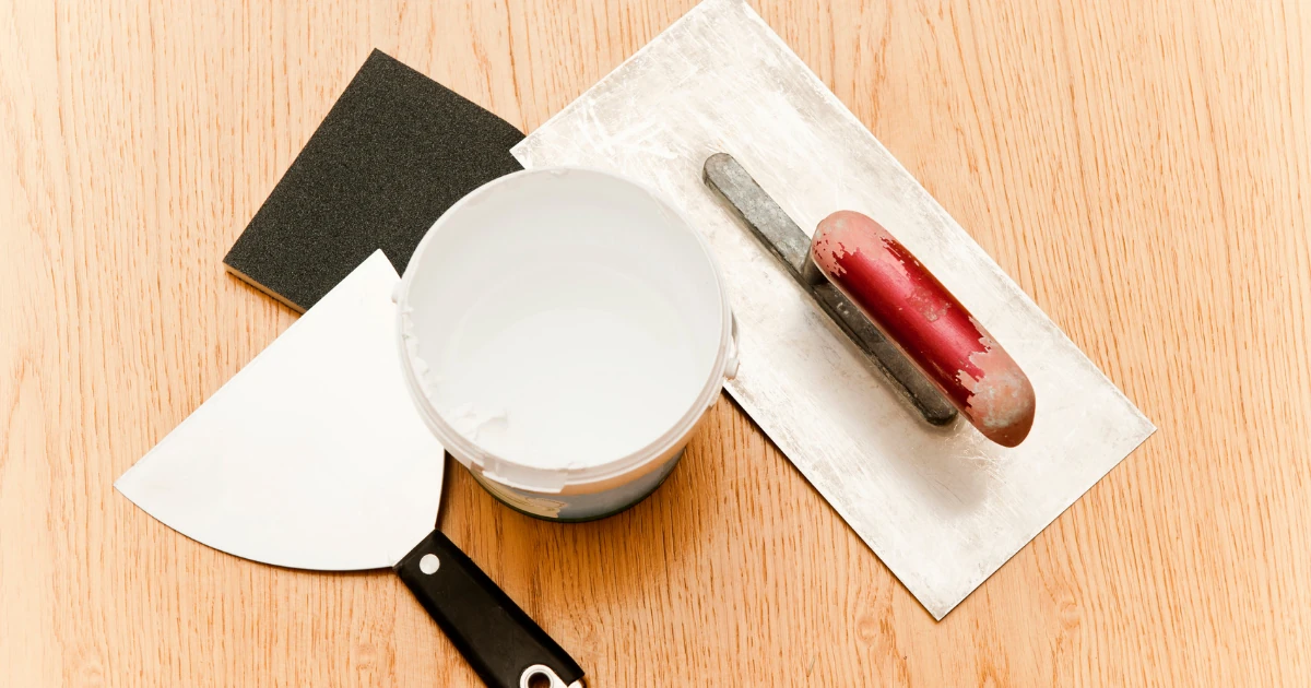 Overhead view of repair tools on a wood surface, including a sanding sponge, putty knife, open tub of white joint compound, and a red plastering trowel on a hawk.