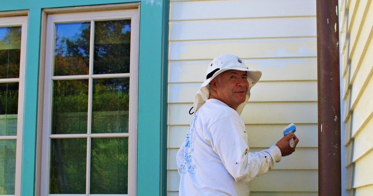A man in a white hat and coveralls paints cream-colored siding with a brush next to a window framed in teal-blue trim.