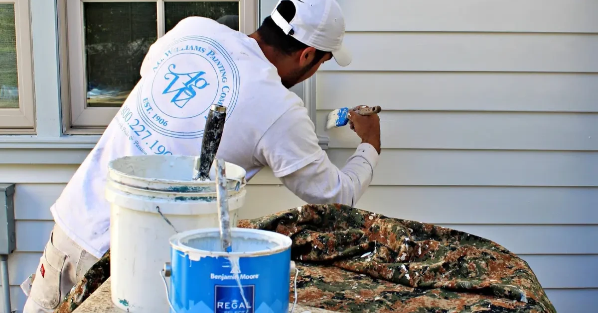 A painter in an A.G. Williams Painting Company shirt applying blue Benjamin Moore Regal Select paint to house siding with a brush, paint buckets in foreground.