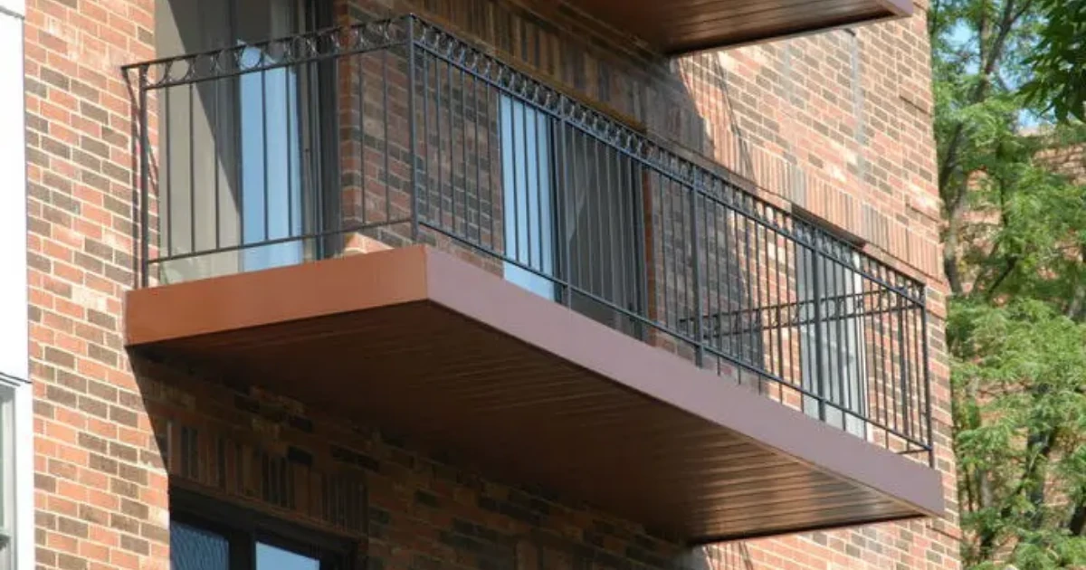 A close-up exterior view of a brick condominium building with a wrought-iron railed balcony and a painted brown balcony underside.