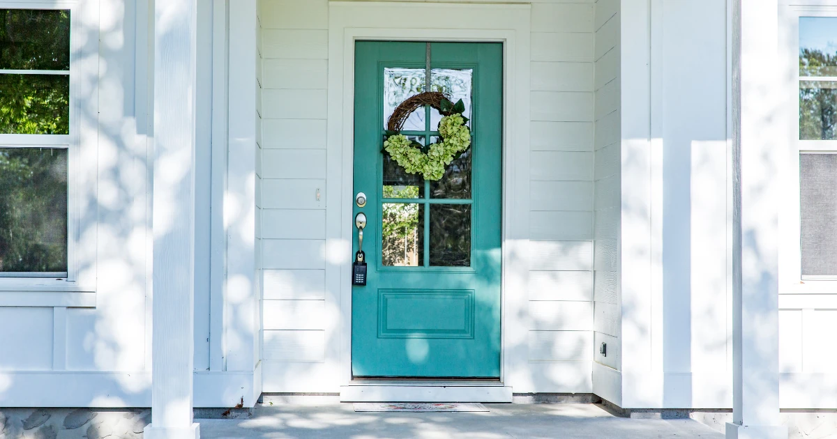 A teal-blue front door with a green hydrangea wreath, set within a white-trimmed entryway on a house with white lap siding.
