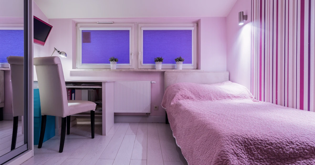 Child's bedroom with a striped pink accent wallpaper wall, a bed with a pink coverlet, a white desk and chair, and two windows with blue shades.