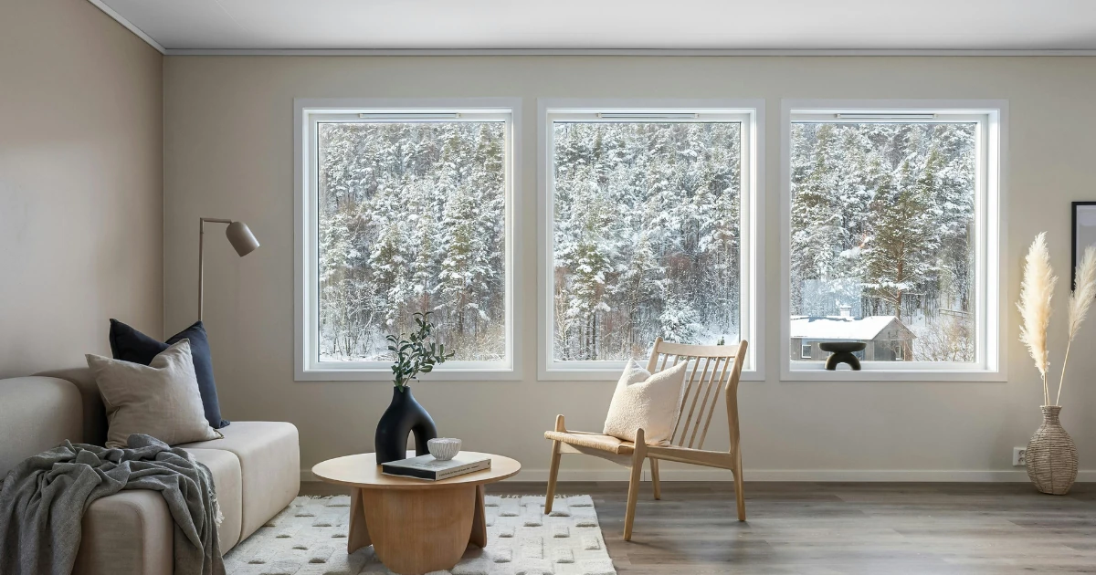 A minimalist living room with greige walls, a beige sofa, and a wood spindle chair, with three large windows framing a snowy forest view.