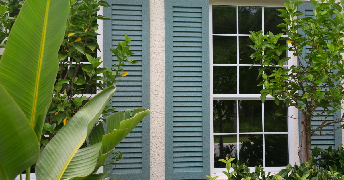 A window with white grille panes flanked by teal-green louvered shutters on a cream stucco exterior, framed by large tropical leaves.