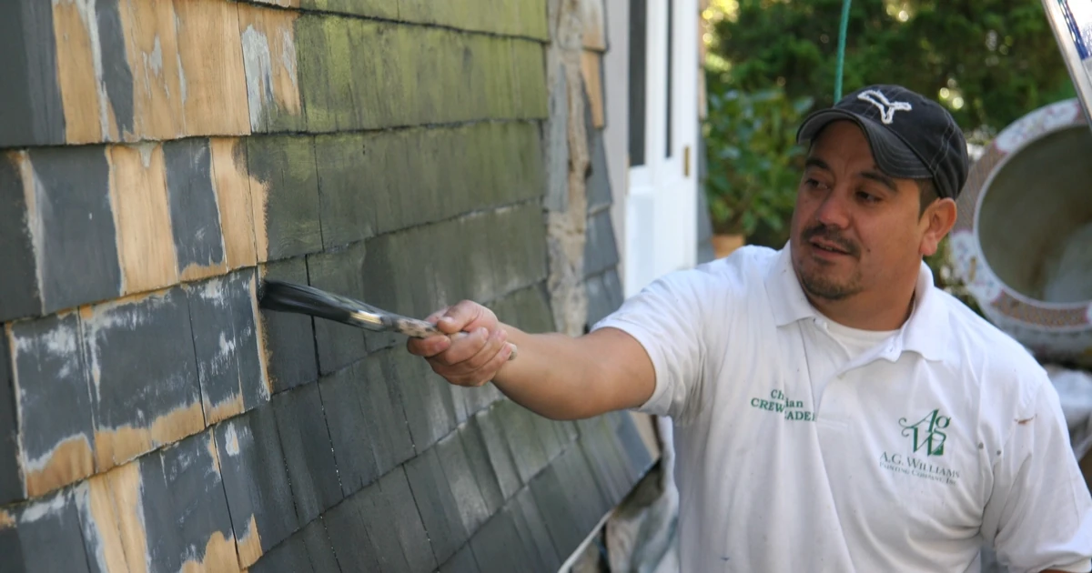 A worker in an A.G. Williams shirt and cap using a handheld brush to paint weathered cedar shingle siding, with bare wood, gray, and green painted sections visible.