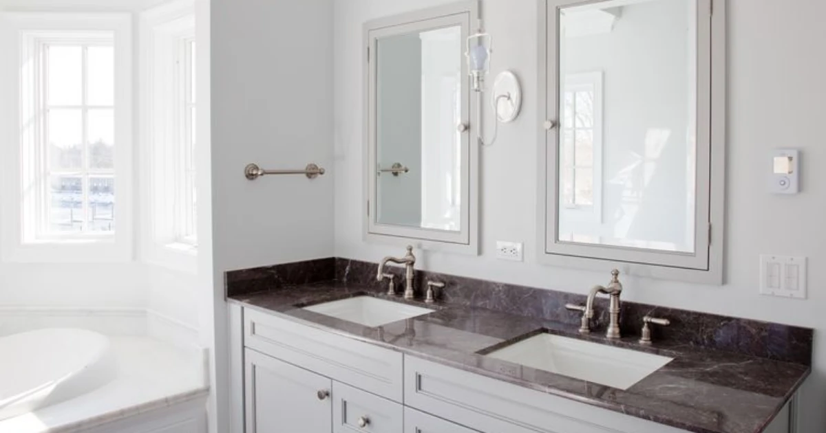 A bright white bathroom with double vanity sinks under dark marble-look countertop, framed mirrors, a wall sconce, and a freestanding tub visible near a window.