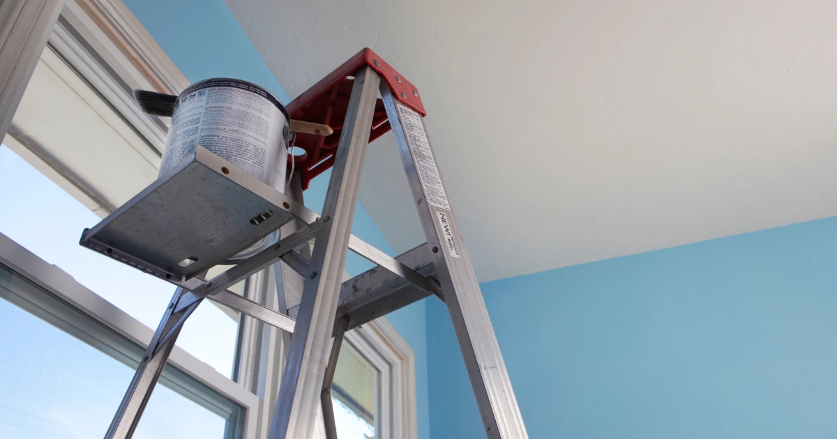 An aluminum stepladder holding a paint can on its tray stands beside a window against a light-blue wall and white ceiling.