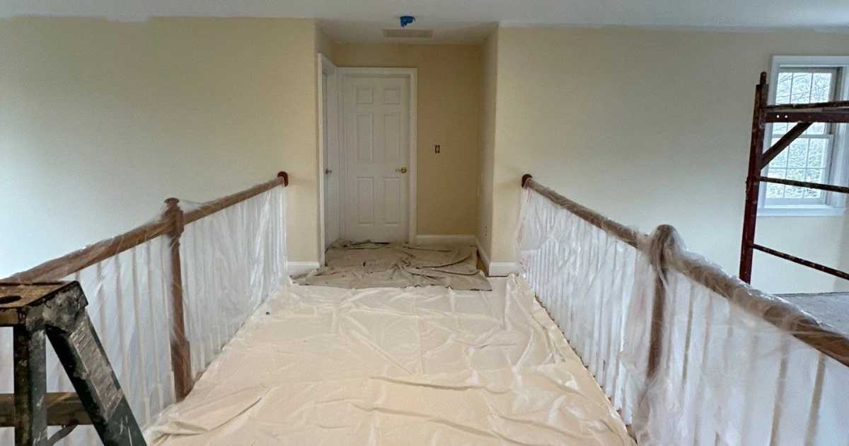 Upstairs hallway prepped for painting with drop cloths on the floor, plastic wrap around the balustrade, a ladder in the corner, and scaffolding by a window.