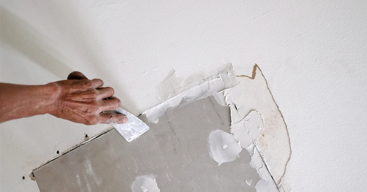 A hand uses a putty knife to spread white joint compound over an exposed drywall patch on a painted wall.