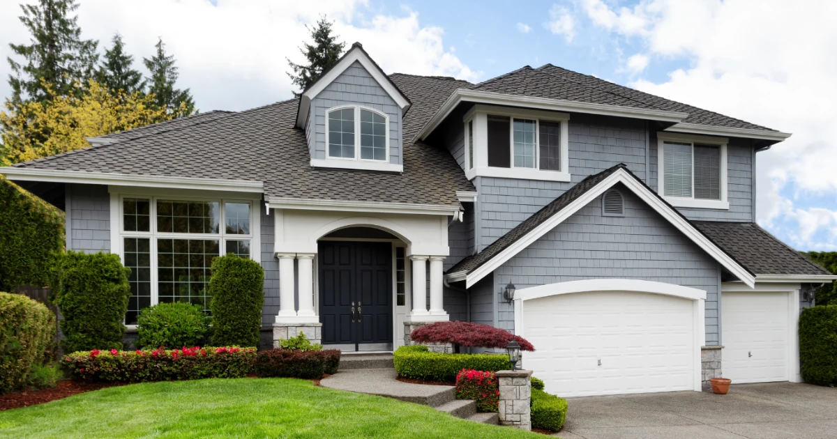 Two-story gray shingle-style house exterior with white trim, a dark front door, stone accents, a three-car garage, and landscaped front yard.