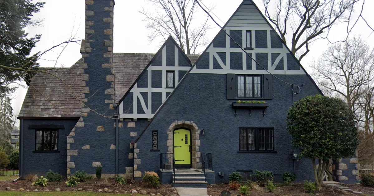 A dark blue-gray Tudor-style house with white half-timber trim and a neon-green front door set in a stone archway.