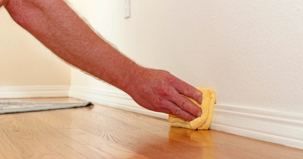 Close-up of a hand wiping a white baseboard with a yellow microfiber cloth next to a hardwood floor.
