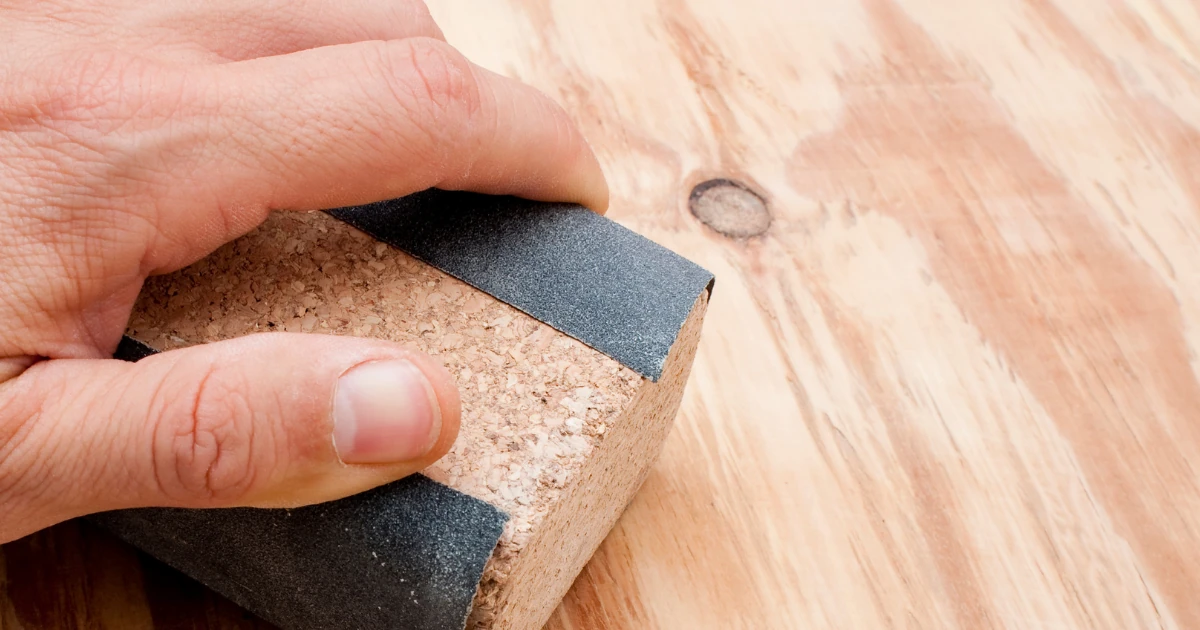 A hand smooths a bare plywood surface using a cork sanding block wrapped in sandpaper, preparing the raw wood for finishing.