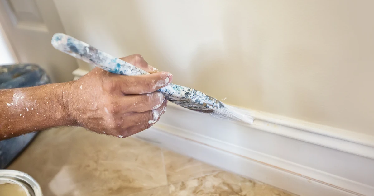 A hand holds a paint-loaded brush, painting a white baseboard along a tiled floor.