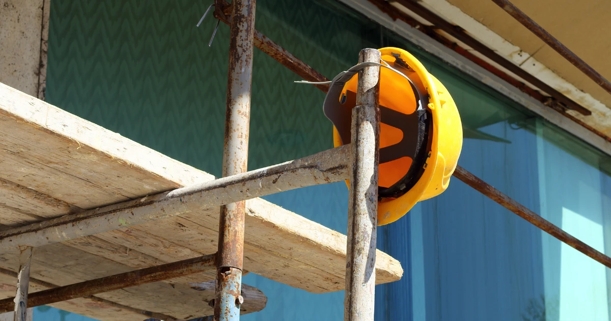 A yellow hard hat hangs on a rusty scaffolding pole beside weathered wooden planks in front of a glass building facade.