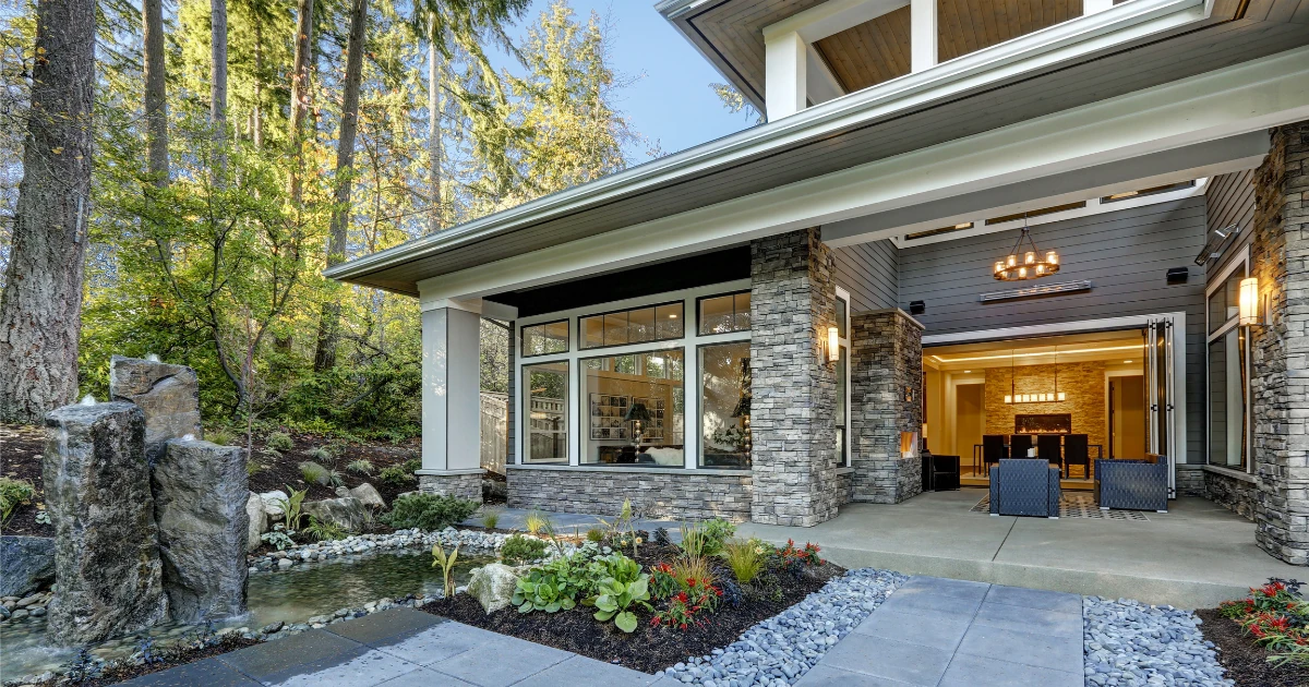 Modern house exterior with stone columns, a covered patio, a small water feature, and an open interior view with a fireplace and dining area visible through large glass doors.