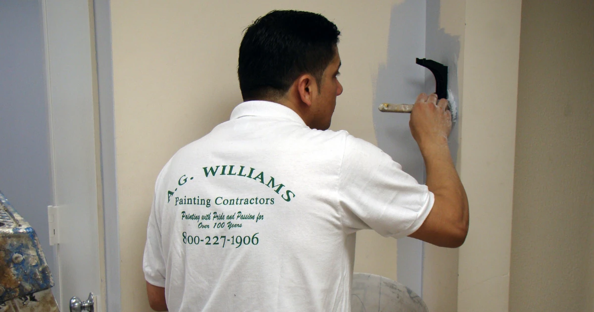 A painter in a white A.G. Williams Painting Contractors shirt brushes light blue paint onto the edge of an interior wall.