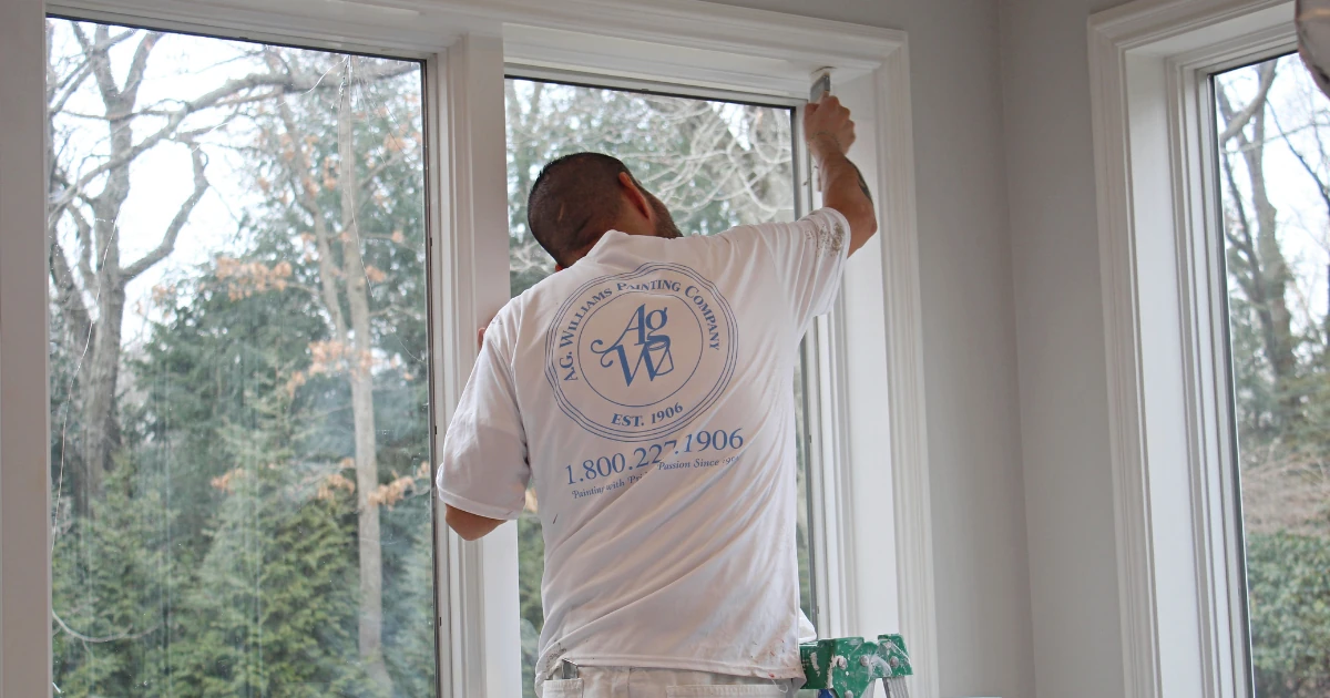 A painter wearing an A.G. Williams Painting Company T-shirt stands on a ladder painting interior window trim in a room overlooking bare trees.