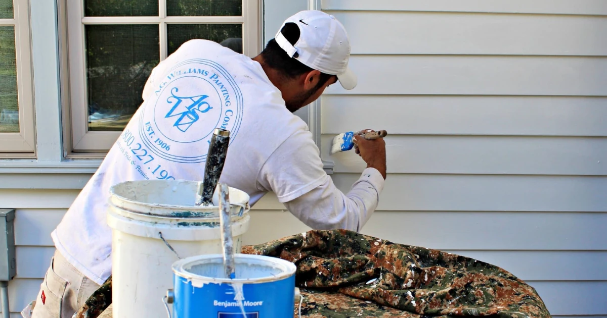 A painter in an A.G. Williams Painting Company shirt and white cap uses a brush to touch up light gray-blue vinyl siding, with paint cans and a drop cloth on a table nearby.