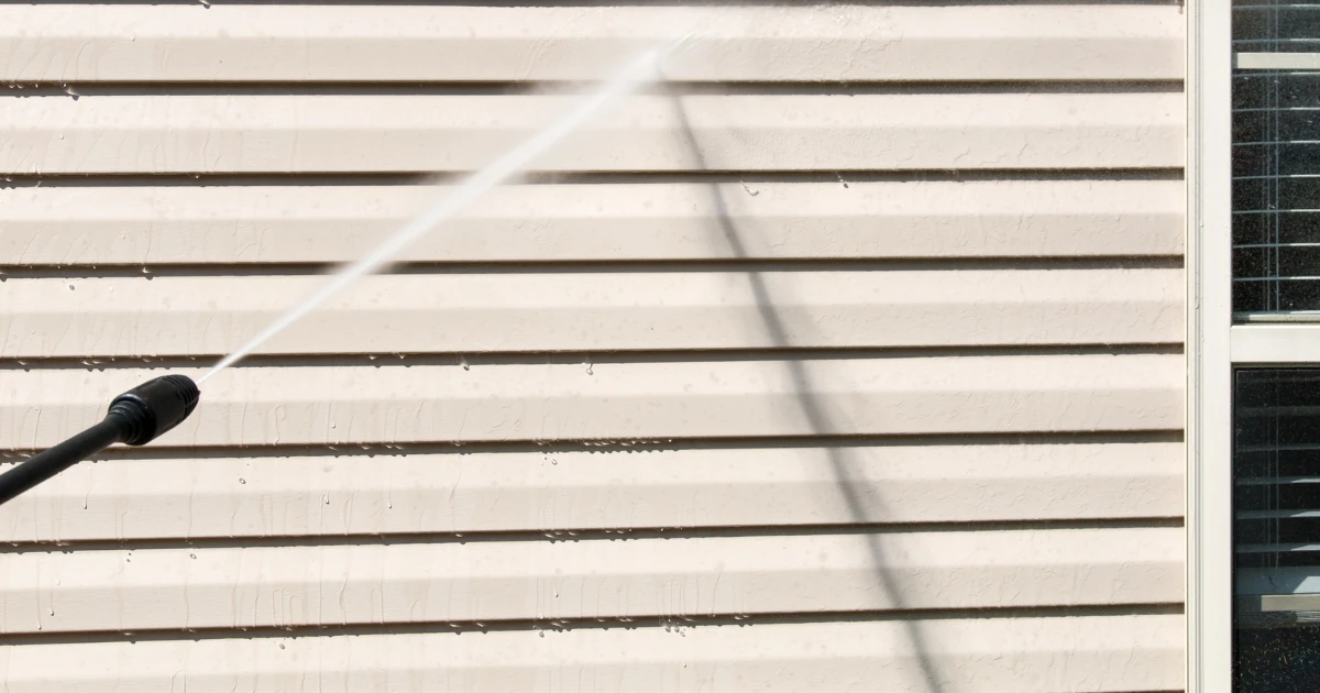 A pressure washer nozzle sprays a stream of water across beige horizontal siding beside a window.