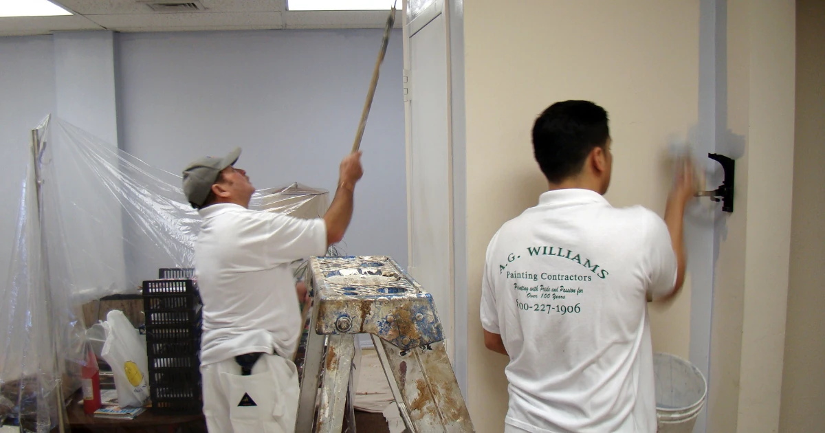 Two painters in white "A.G. Williams Painting Contractors" uniform shirts work in a commercial interior, one rolling a ceiling edge from beside a ladder.