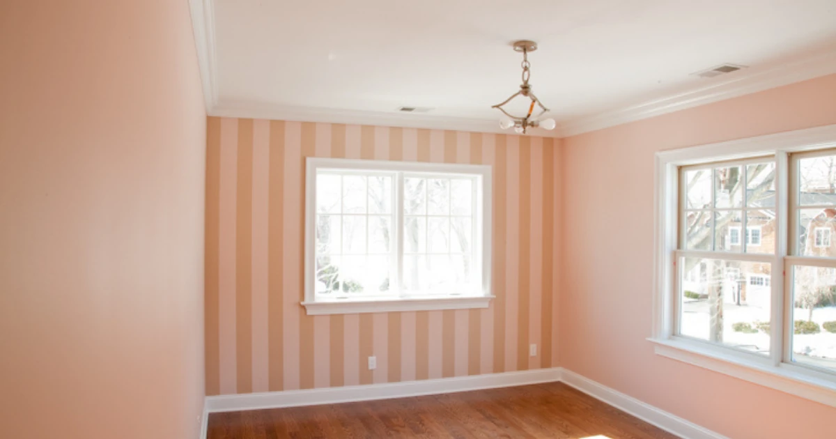 An empty room with a pink and tan vertically striped wallpaper accent wall, white crown molding, a hardwood floor, and two large windows.