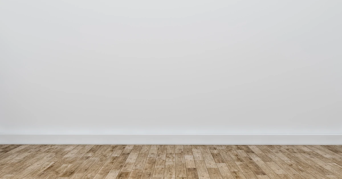 Plain white painted wall with matching white baseboard above a light hardwood plank floor, with no furnishings in view.