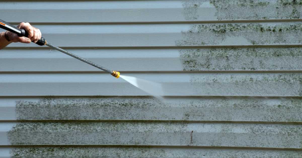 A hand holds a pressure washer wand, spraying vinyl siding that is clean on the left and covered in green mildew on the right.