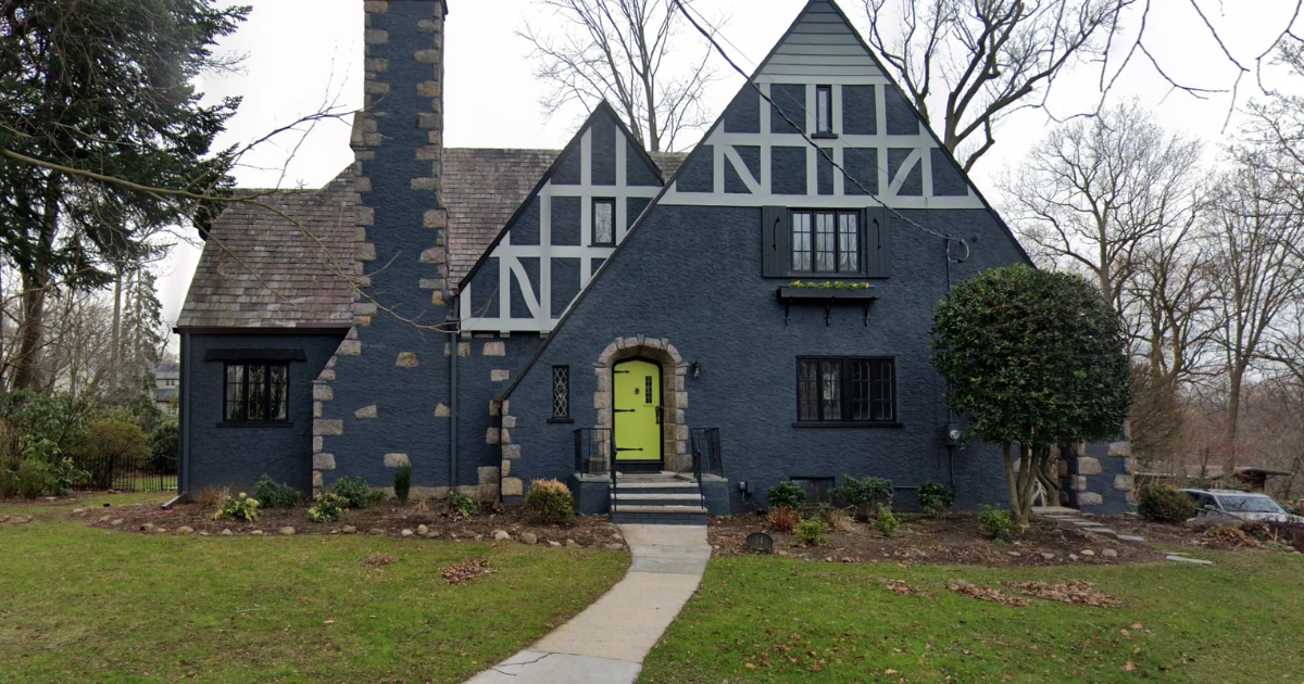 Tudor-style stone-and-stucco house painted dark blue with light gray half-timbering and a bright chartreuse front door, viewed from the front lawn.