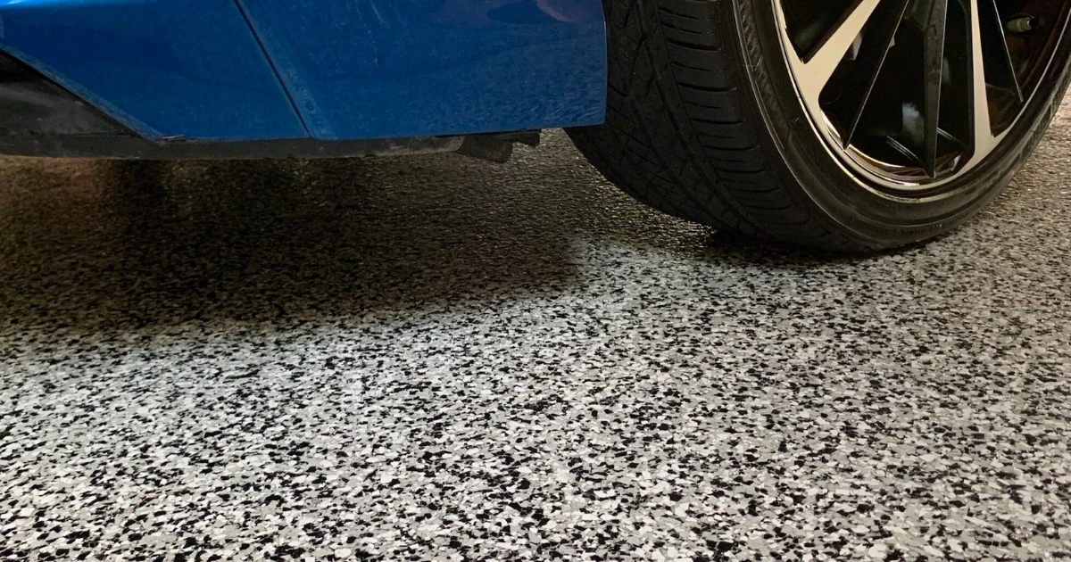 Close-up of a blue car's tire and lower body parked on a black, white, and gray flake epoxy floor.