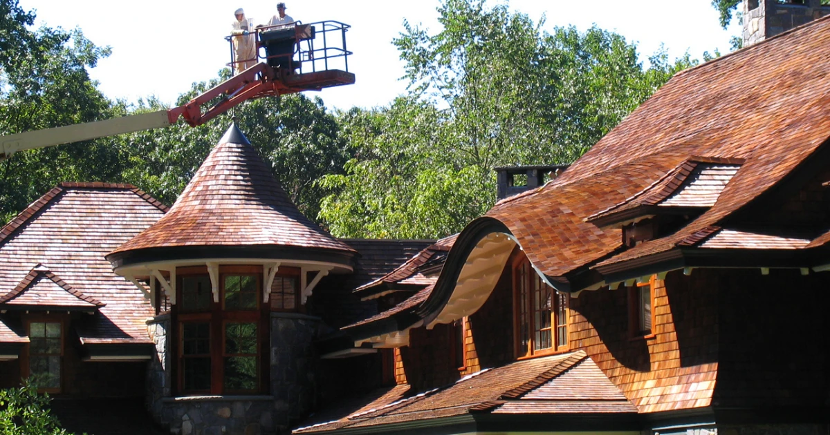 Two workers in an orange boom lift work above the ornate cedar-shingle roofline and turret of a shingle-style home surrounded by trees.