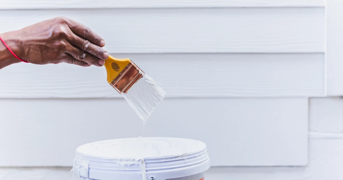 A close-up of a hand dipping a paintbrush with copper ferrule into white paint above a white paint bucket, set against white vinyl siding.