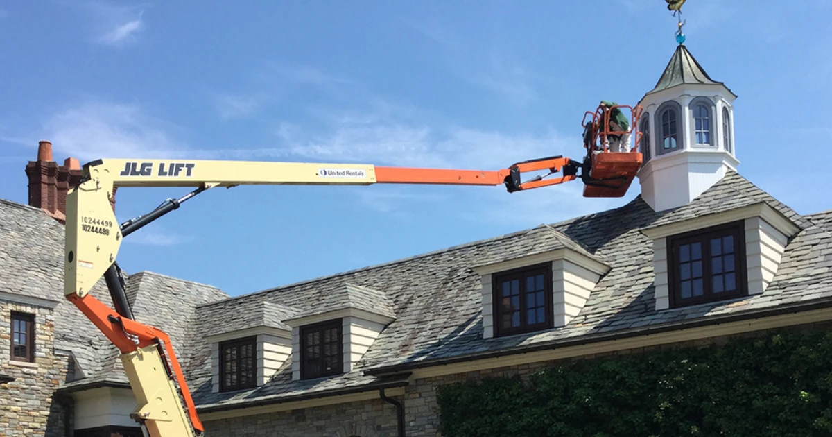 A worker in an orange boom lift basket reaches a white cupola atop the slate roof of a stone house against a clear blue sky.