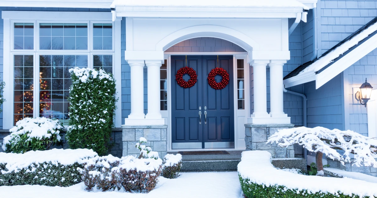 Snow-covered front entrance of a blue-gray shingled home with white columns and double navy doors hung with two red berry wreaths.