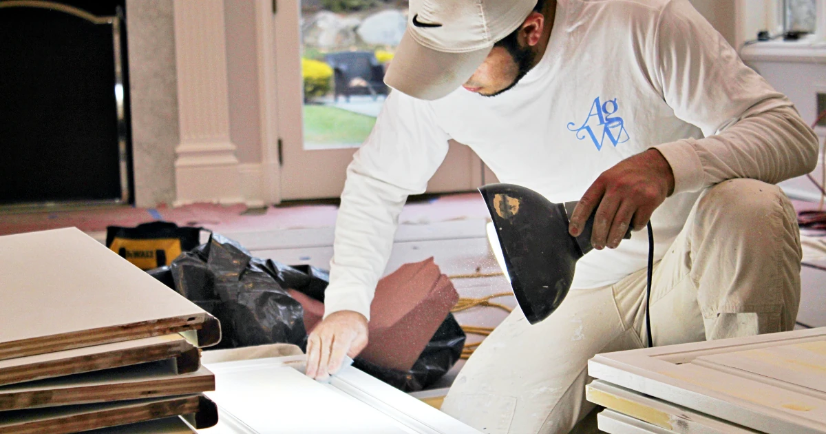 A crew member in a white long-sleeve shirt with a blue AGW logo and a Nike cap sands white cabinet doors laid out inside a home.