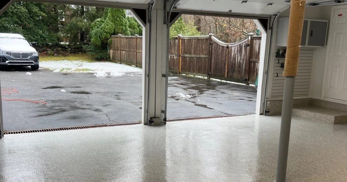 The interior of a two-car garage with a light speckled epoxy floor, looking out through the open door at a driveway with melting snow and a parked SUV.