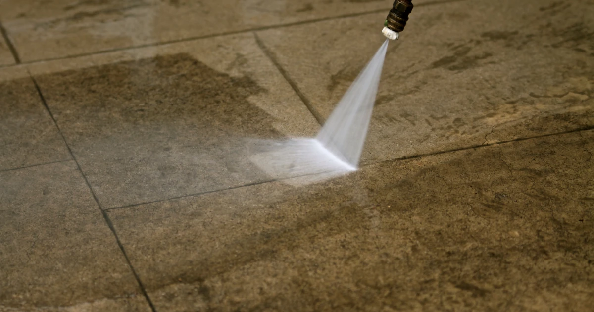 A pressure washer spray cleans a dirty stone paver surface, leaving a visibly lighter cleaned patch across the pavers.