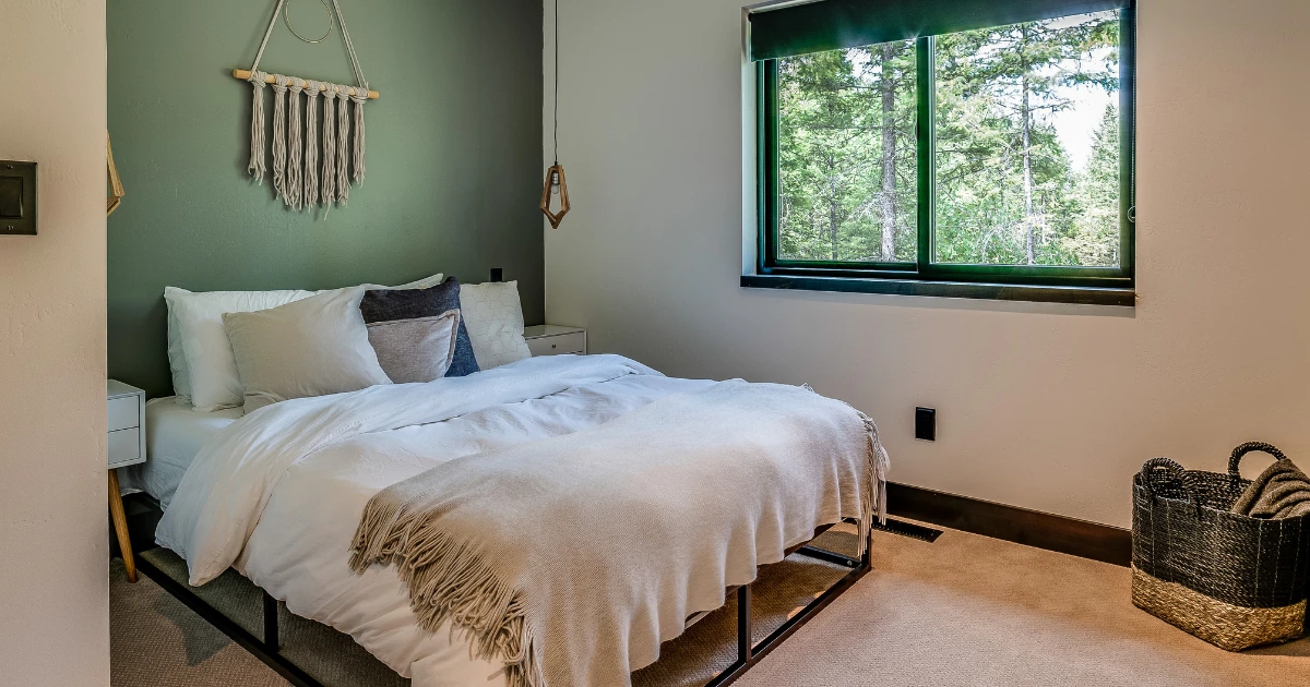 Bedroom with a sage green painted accent wall, a macrame wall hanging over the bed, and a window looking out onto trees.