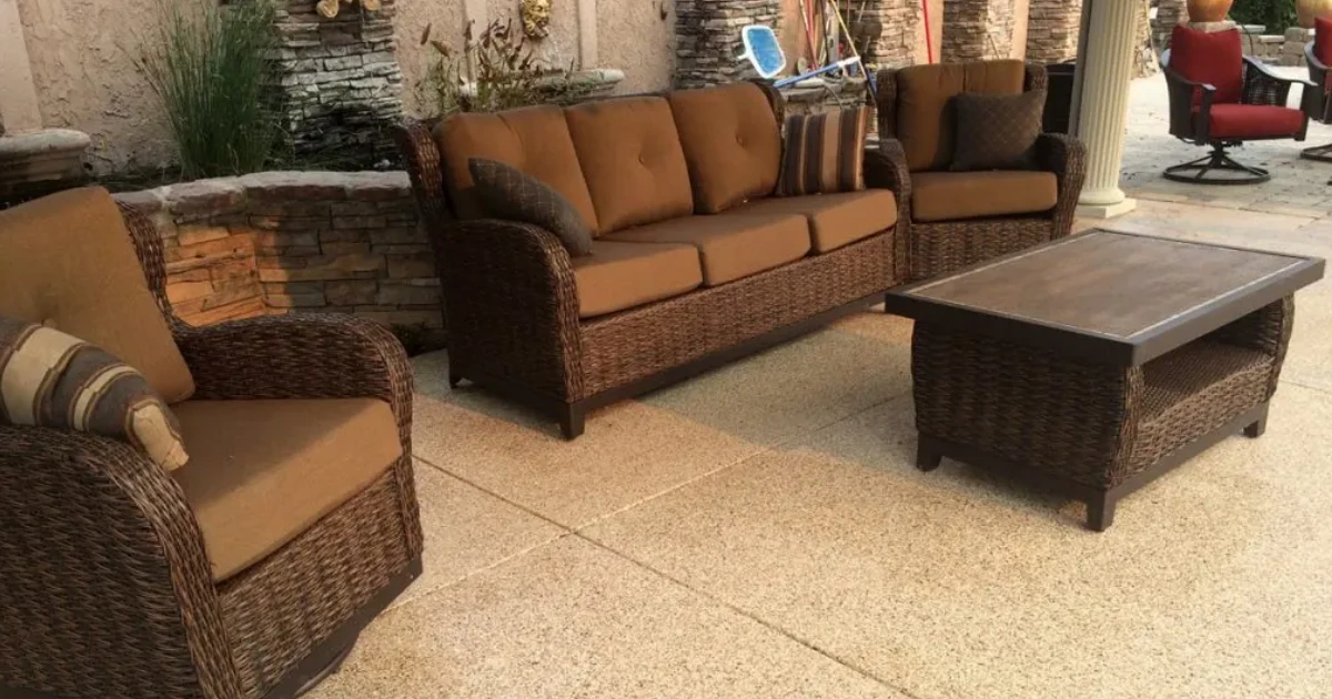 A wicker patio furniture set with brown cushions and a coffee table arranged on a coated concrete patio floor.