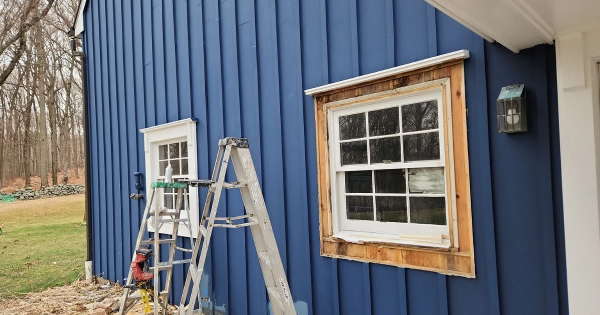 Exterior of a house with dark blue vertical siding under renovation, showing two windows with exposed raw wood trim awaiting paint and ladders set up nearby.