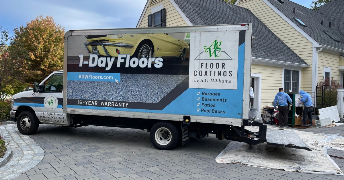 An A.G. Williams branded box truck advertising "1-Day Floors" and "Floor Coatings by A.G. Williams" parked in a driveway, with two workers in blue AGW hoodies unloading equipment near a house.